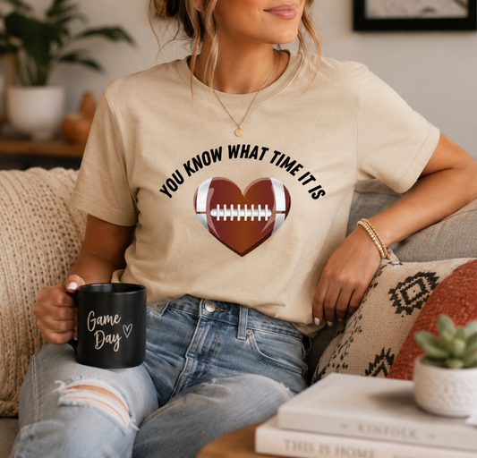 A woman sitting on a couch wearing a beige graphic t-shirt with a football heart design and the text 'YOU KNOW WHAT TIME IT IS'. She is holding a black mug that says 'Game Day' and wearing blue jeans in a cozy living room setting.
